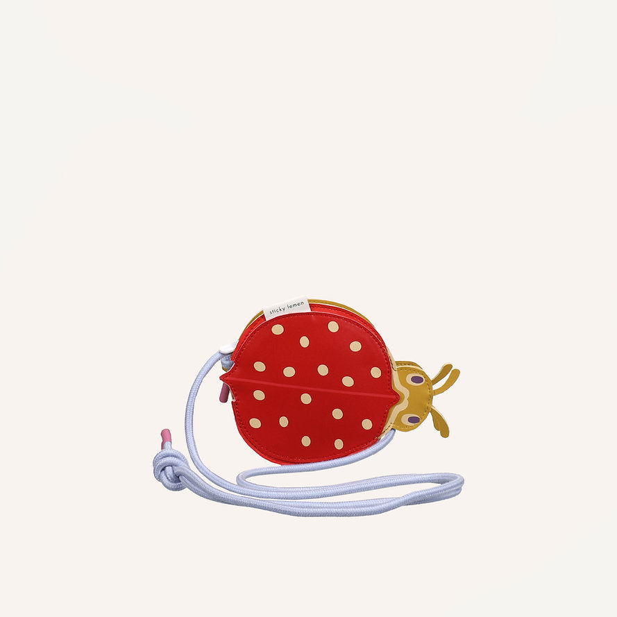 The Sticky Lemon Wallet Bag - Ladybug is a small, round kids bag made of red PU leather with yellow polka dots and a ladybug design. It features a light purple strap and is shown on an off-white background.