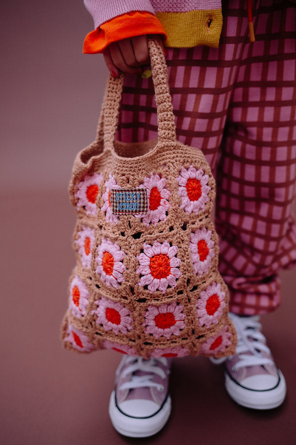 A person wearing checkered pants and sneakers holds the Sticky Lemon Crochet Bag - Lemon Tree - Flower - Campfire Orange, featuring playful pink and red flower squares—perfect as a kids’ accessory.