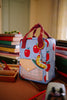 The Sticky Lemon Backpack Small - Special Edition - Apple Grub is a small blue kids’ backpack with red apple prints, a plush caterpillar keychain, waterproof lining, and is made from recycled PET bottles. Seen on a desk with books and school supplies.