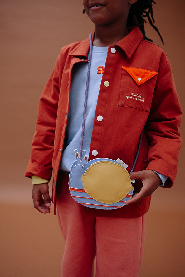 A child in a red jacket with orange accents and pink pants holds the Sticky Lemon Wallet Bag - Snail, set against a plain warm brown background.