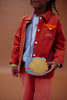 A child in a red jacket with orange accents and pink pants holds the Sticky Lemon Wallet Bag - Snail, set against a plain warm brown background.