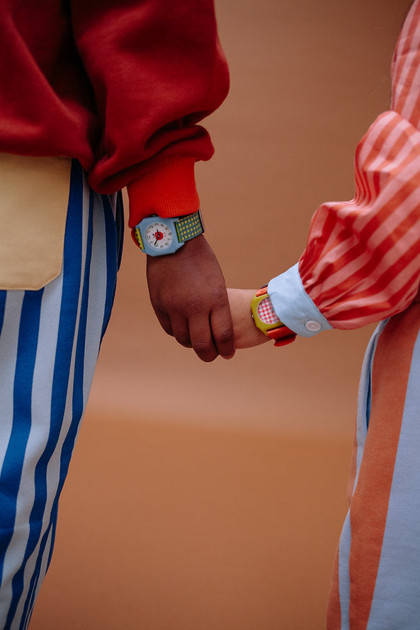 Two people hold hands—one in a red sweater and blue-and-white striped pants, the other in a red-and-white striped shirt. Both wear Sticky Lemon x Mini Kyomo Watch - Apple, adding pops of color to their wrists. The warm background is softly blurred.