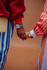 Two people hold hands—one in a red sweater and blue-and-white striped pants, the other in a red-and-white striped shirt. Both wear Sticky Lemon x Mini Kyomo Watch - Apple, adding pops of color to their wrists. The warm background is softly blurred.