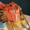 The Sticky Lemon Backpack Large | Color Blocking in Coral Pink + Reed Green, featuring orange handles and a green strap, sits on a tiled table by a lemon, milk carton, and bread slices. Its main pocket is open to show the waterproof lining and white zipper.