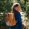A young girl with long dark hair, wearing a blue sweater and carrying the Sticky Lemon Backpack Small Special Edition | Adventure Collection | Khaki Green by its shoulder handle, smiles on a sunlit path in a green forest.