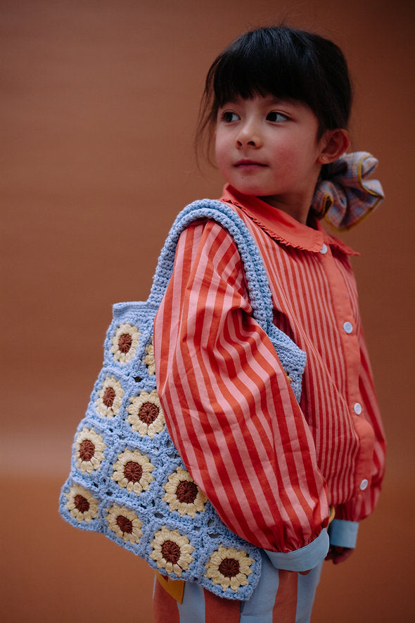 A young child in a red and orange striped shirt holds the Sticky Lemon Crochet Bag - Lemon Tree - Flower - Willow Basket over their shoulder, standing against a plain brown background.