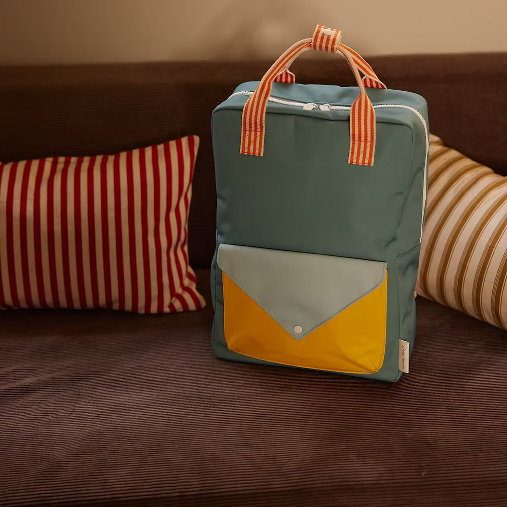 The Sticky Lemon Backpack Large | Envelope | Diving Suit, made from recycled PET with a waterproof lining, features blue with orange striped handles and yellow/light blue pocket. It sits on a brown sofa with red/white and beige/white striped cushions.