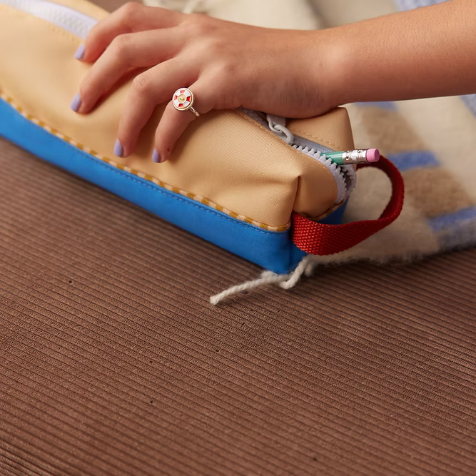 A hand with purple nail polish and a colorful ring holds the Sticky Lemon Pencil Case | Color Blocking | Sandy Yellow + Radar Blue, featuring a red strap and partially unzipped to reveal a pencil and pen inside.
