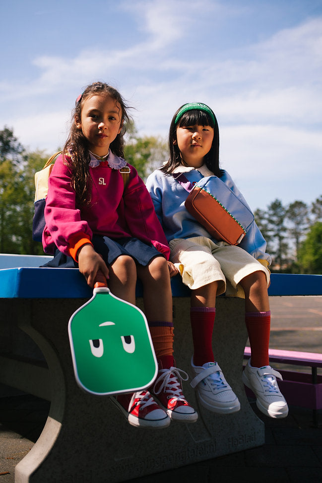 Two children sit side-by-side on a bench outdoors. One wears a pink sweatshirt and navy skirt, holding a green paddle, while the other dons a light blue top and yellow shorts. Both have backpacks and wear sneakers. A Sticky Lemon Pick Ball Game Set lies next to them, with trees visible in the background under a clear sky.