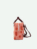 A small, coral pink Sticky Lemon backpack with a subtle apple print, featuring a front zipper pocket and adjustable brown straps, set against a plain white background.