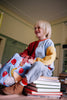 A blond-haired, smiling child sits on books wearing a colorful sweatshirt and boots, holding the Sticky Lemon Backpack Small – Special Edition – Apple Grub, made from recycled PET bottles with red apple prints and a waterproof lining.