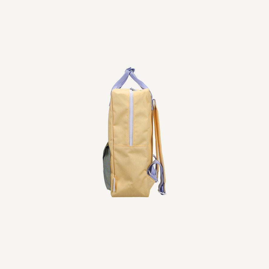Side view of the Sticky Lemon Backpack Large - Envelope in Twiggy Beige, a light yellow kids' backpack with lavender handles and zippers, orange straps, mesh side pocket, made from recycled PET bottles on a plain white background.