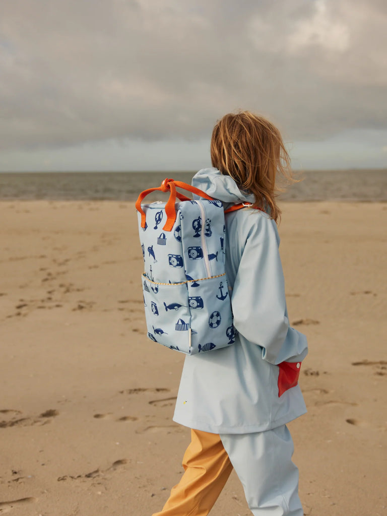 A person in a light blue raincoat and yellow pants walks on a sandy beach, carrying a Sticky Lemon Backpack Large - Special Edition - Wonders of the Deep Sea. The sky is cloudy and the sea can be seen in the background.