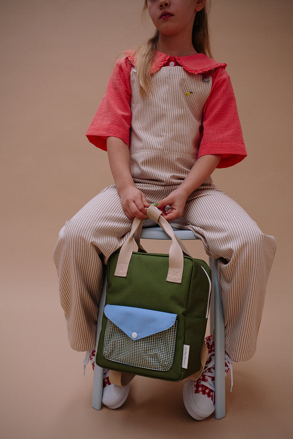 A young child in a red-collared shirt and striped overalls sits on a chair, holding the Sticky Lemon Backpack Small - Envelope - Tickling Grass, which is waterproof, made from recycled PET bottles, and features beige handles and a blue front pocket.