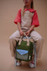 A young child in a red-collared shirt and striped overalls sits on a chair, holding the Sticky Lemon Backpack Small - Envelope - Tickling Grass, which is waterproof, made from recycled PET bottles, and features beige handles and a blue front pocket.