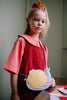 A young girl in a red dress with a large collar stands indoors, wearing a yellow Sticky Lemon Wallet Bag - Snail and a red scrunchie, looking slightly to the side with a neutral expression.