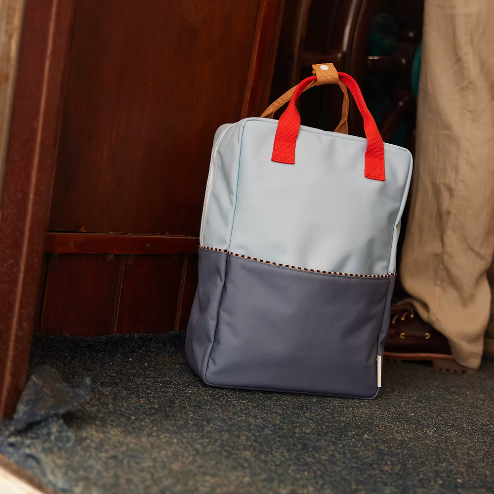 The Sticky Lemon Backpack Large | Color Blocking | Bissou Blue + Blue Whale, crafted from recycled PET bottles, features a light blue top, dark blue bottom, and red handles, shown on a carpet floor beside a wooden wall and pant leg.