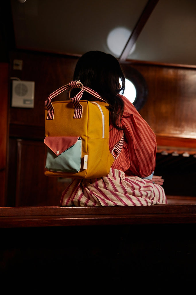 A person with long dark hair, in a red and white striped outfit, sits on a wooden bench with their back to the camera, carrying the Sticky Lemon Backpack Small | Envelope Collection | Submarine Yellow.