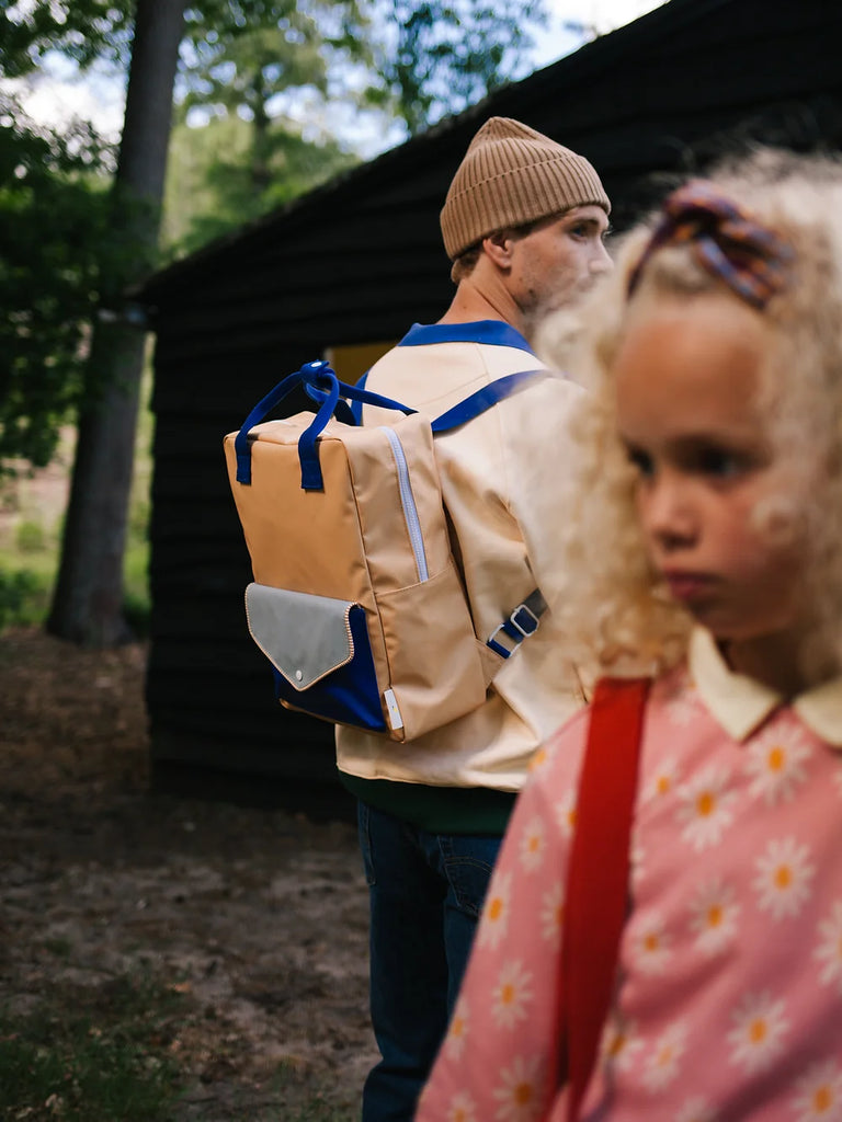 A young girl in a floral dress appears slightly out of focus in the foreground, while a man wearing a beanie and carrying a Sticky Lemon Backpack Large from the Envelope Collection in Camp Yellow stands sharply focused in the background.