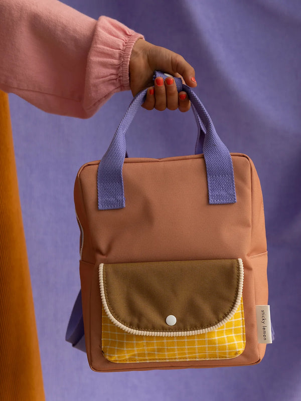 A person holding a Sticky Lemon Backpack Small in Harvest Moon, made from recycled PET bottles, with a yellow plaid pocket and a purple handle against a lavender background.