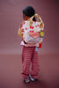 A young child with long dark hair stands facing away, wearing the Sticky Lemon Backpack Small - Special Edition - Snail Mail, made from recycled PET bottles, and loose checked red pants against a plain brown backdrop.