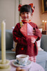 A young girl with dark hair in pigtails and a red bow stands indoors, smiling as she holds the Sticky Lemon Fanny Pack Small – Envelope – Rosebay Pink. She wears a red sweater with orange dots and patterned pants. A table with a teacup and candle is nearby.
