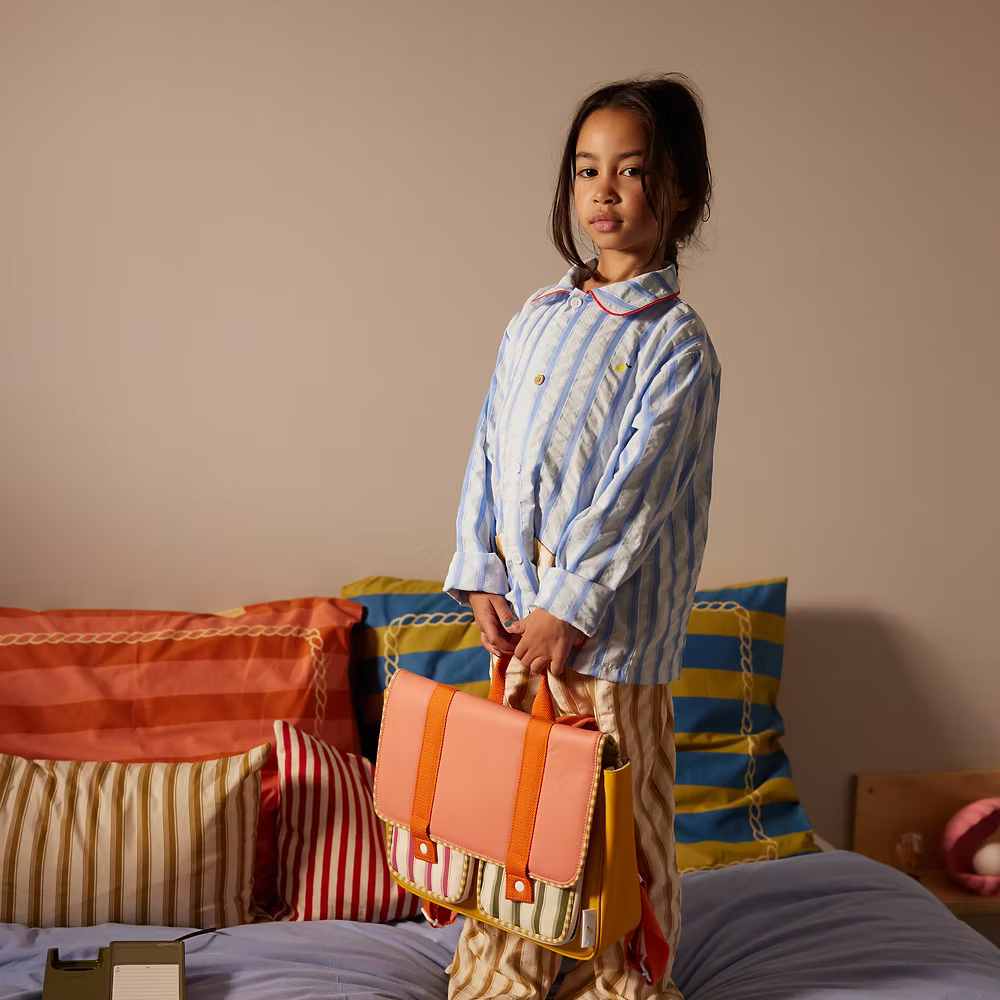 A young girl in striped pajamas stands on a bed with colorful striped pillows, holding the Sticky Lemon School Bag in Coral Pink + Submarine Yellow. The background is a plain wall.