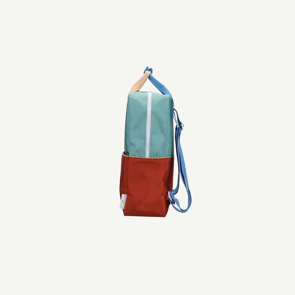 Side view of the Sticky Lemon Backpack Large | Color Blocking | Aquatic Green + Shipwreck Rust, made from recycled PET, featuring a teal upper, red lower section, blue adjustable strap, tan top handle, and waterproof lining on a white background.