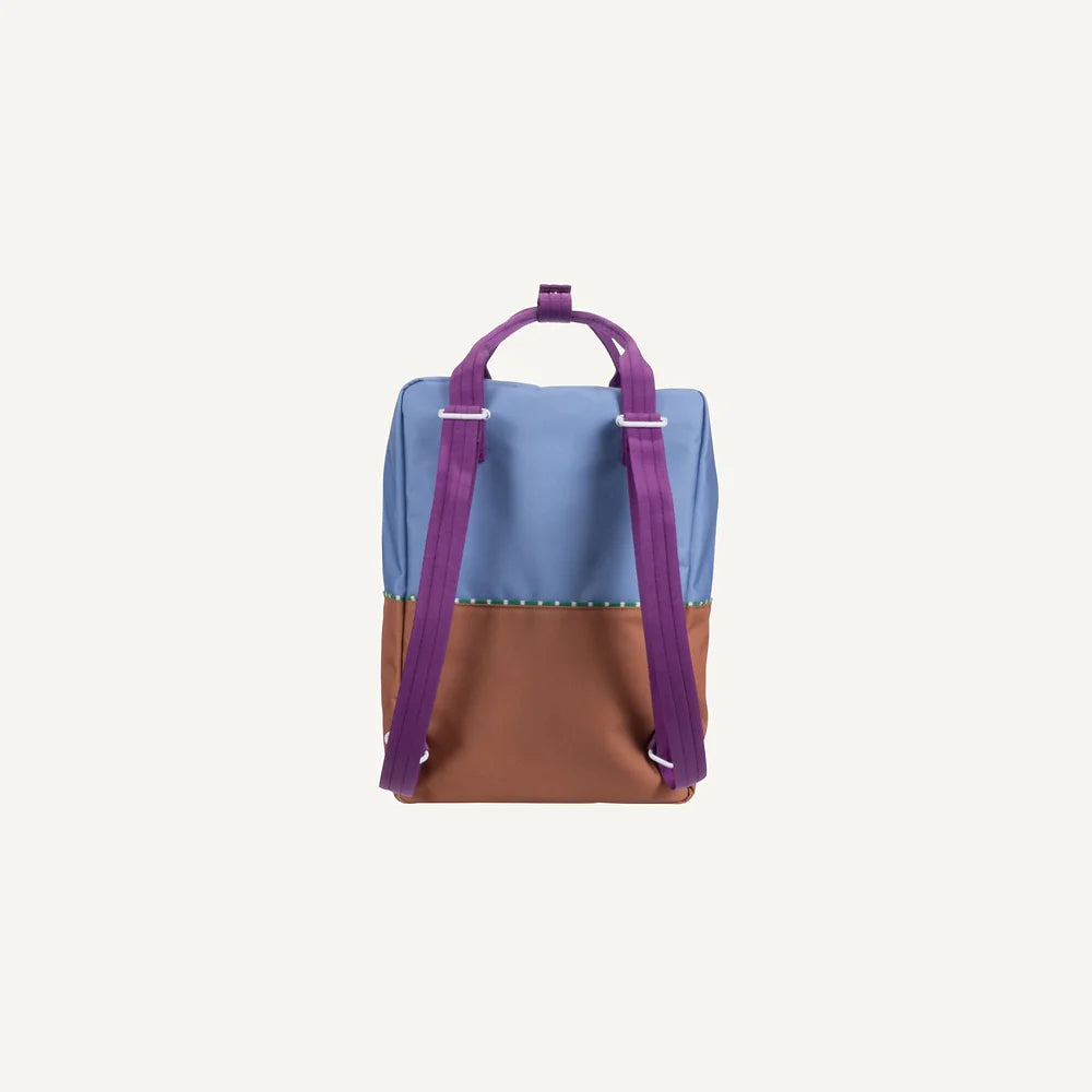 The Sticky Lemon Backpack Large in the Color Blocking edition showcases a vibrant design with a spacious blue top section and brown bottom section. It comes equipped with purple adjustable shoulder straps and a coordinating purple carrying handle. Crafted from recycled PET bottles, this backpack boasts a minimalistic style and is displayed on a plain off-white background.