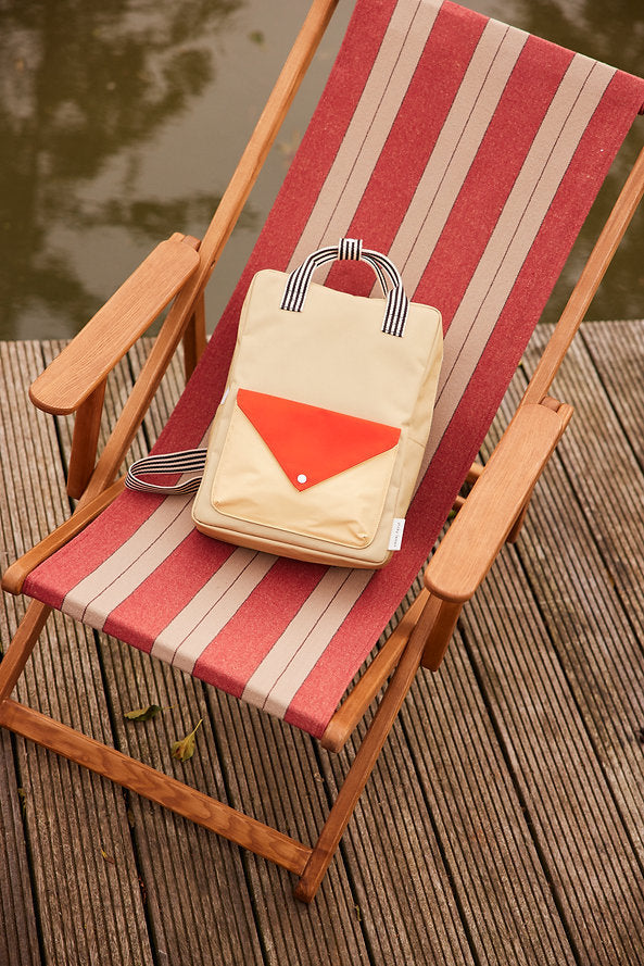 A Reed Green Sticky Lemon Backpack Large - Envelope, crafted from recycled PET bag material and featuring striped handles in beige and red, rests on a red-and-beige striped deck chair placed on a wooden deck by the water.