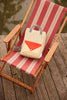 A Reed Green Sticky Lemon Backpack Large - Envelope, crafted from recycled PET bag material and featuring striped handles in beige and red, rests on a red-and-beige striped deck chair placed on a wooden deck by the water.