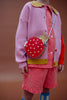 A child wears a pink knit cardigan with yellow accents over a red gingham shirt and shorts, accessorized with colorful nails and the Sticky Lemon Wallet Bag - Ladybug. The playful accessories pop against the plain brown background.
