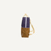 Side view of a vertically oriented Sticky Lemon Backpack Large with a color-block design titled Skate Ramp + Golden Cup. The top half is navy blue, while the lower half is brown. The backpack features beige straps and a white vertical stripe in the middle. It boasts adjustable shoulder straps and a minimalist, rectangular shape.