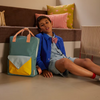A young boy in a blue jacket and bow tie sits on the floor by a bench with patterned pillows. Next to him is the Sticky Lemon Backpack Large | Envelope | Diving Suit, featuring yellow, blue, and orange accents, made from recycled PET bottles with waterproof lining.