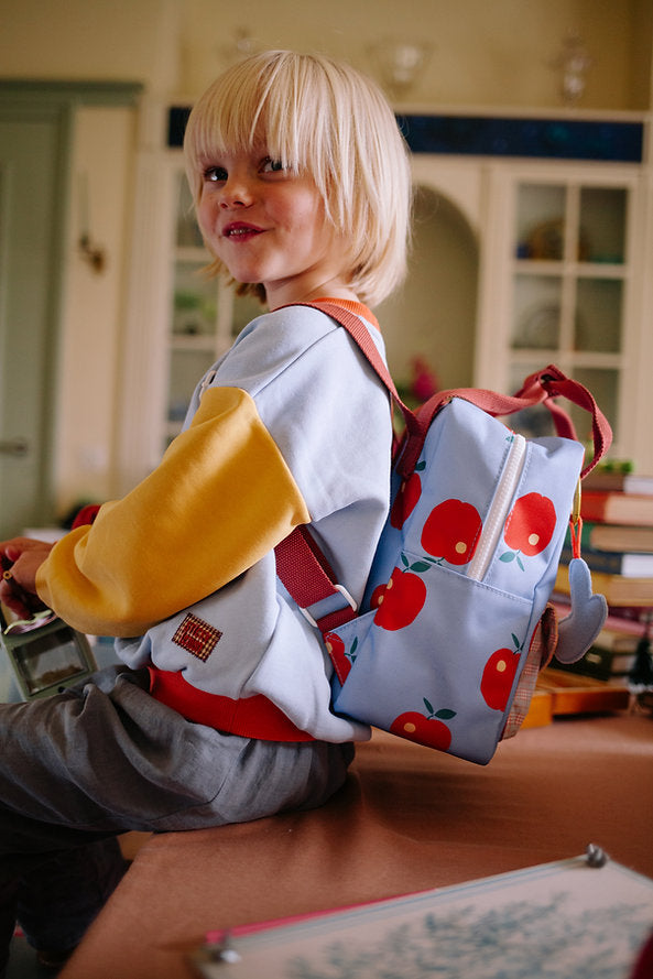 A young blonde child wearing a colorful sweatshirt and gray pants sits on a table, smiling. They carry the Sticky Lemon Backpack Small - Special Edition - Apple Grub, made from recycled PET bottles with a red apple pattern and waterproof lining.
