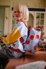 A young blonde child wearing a colorful sweatshirt and gray pants sits on a table, smiling. They carry the Sticky Lemon Backpack Small - Special Edition - Apple Grub, made from recycled PET bottles with a red apple pattern and waterproof lining.