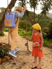 A boy wearing beige pants and blue sneakers swings on a rope swing, while a girl in an orange Sticky Lemon Backpack Large dress and yellow hat looks up at him, standing on a gravel path surrounded by greenery.