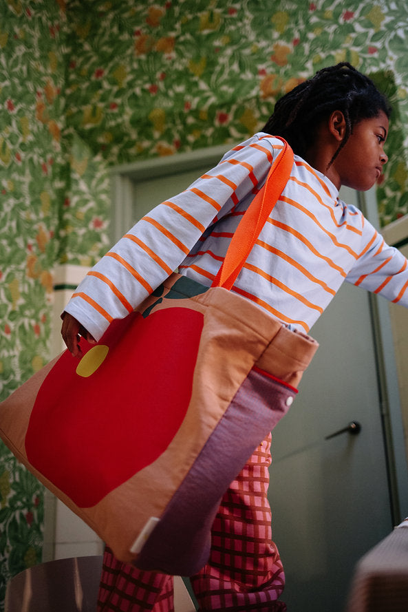 A child in a striped shirt and checkered pants holds the Sticky Lemon Shopper - Apple tote, featuring a large red circle and yellow dot, while standing in a room with green floral wallpaper.