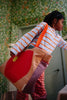 A child in a striped shirt and checkered pants holds the Sticky Lemon Shopper - Apple tote, featuring a large red circle and yellow dot, while standing in a room with green floral wallpaper.