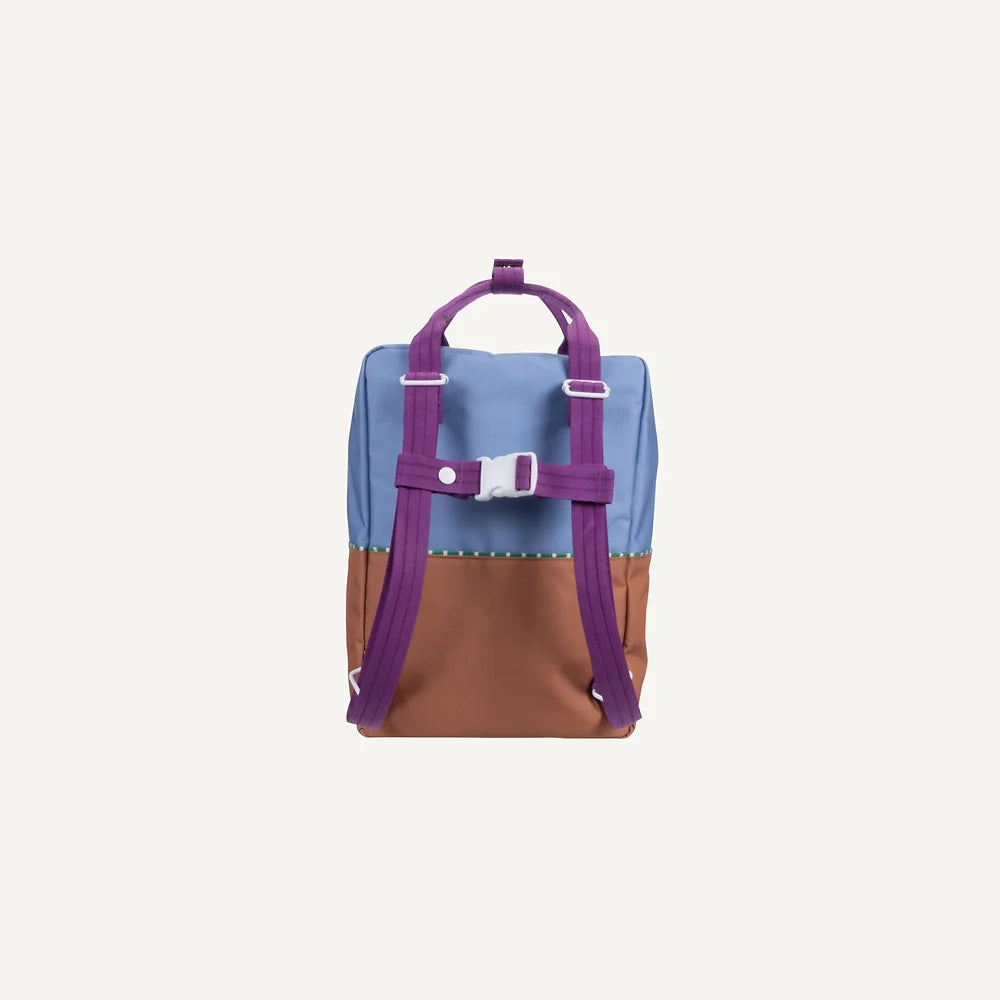 A rectangular Sticky Lemon Backpack Large is shown from the back. It features a color-block design with a badminton blue top section, a leather ball brown bottom section, and purple adjustable shoulder straps and handle. The straps are connected with a white buckle for extra support. The background is plain white.