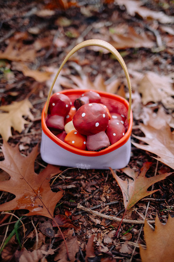 The Sticky Lemon Treasure Bag - Dewy Lilac rests among fallen brown leaves and twigs in a forest, filled with red and white spotted toy mushrooms.