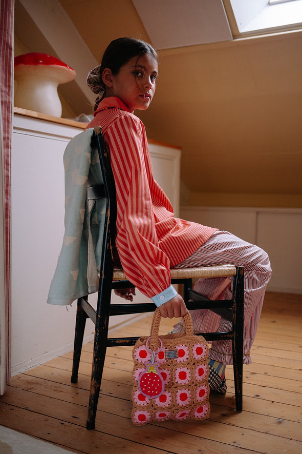 A girl in striped pajamas sits sideways on a chair, looking back over her shoulder. She holds a charming Sticky Lemon Crochet Bag - Lemon Tree - Flower - Campfire Orange, and a jacket is draped over the chair. The room has wooden floors and warm lighting.