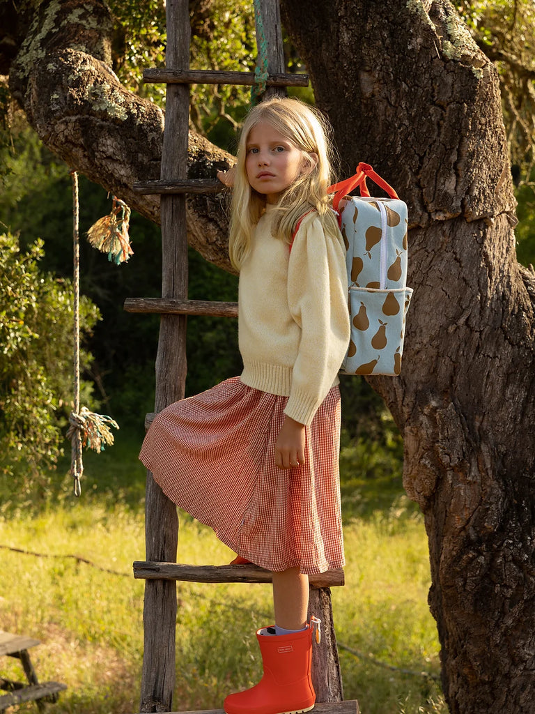 A young girl in a white sweater, coral skirt, and red boots stands on a wooden ladder against a large tree with a Sticky Lemon Backpack Large featuring a YKK zipper hanging on one side.