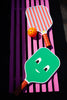A pink bench holds a Sticky Lemon Pick Ball Game Set. One paddle is green with a smiling face, while the other has red and pink stripes. Both paddles have orange handles, and the ball is perforated with small holes, completing this colorful set.
