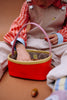 A child in striped overalls and a red, white, and blue shirt reaches into their Sticky Lemon Treasure Bag - Campfire Orange with a pink handle, seated on a brown surface. An acorn and a striped cloth lie nearby.
