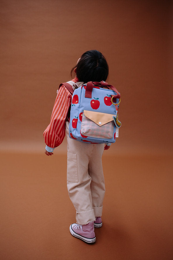 A young child faces away, wearing the Sticky Lemon Backpack Small - Special Edition - Apple Grub, made from recycled PET bottles with waterproof lining and red apple prints, plus a red-orange striped top and beige corduroy pants against a brown background.