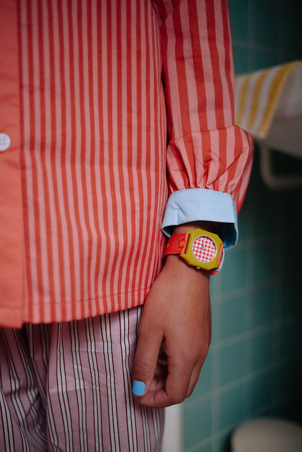 A person in pink striped pajamas and a Sticky Lemon x Mini Kyomo Watch - Flower with a checkered face stands in a tiled bathroom. Their fingernails are painted light blue.