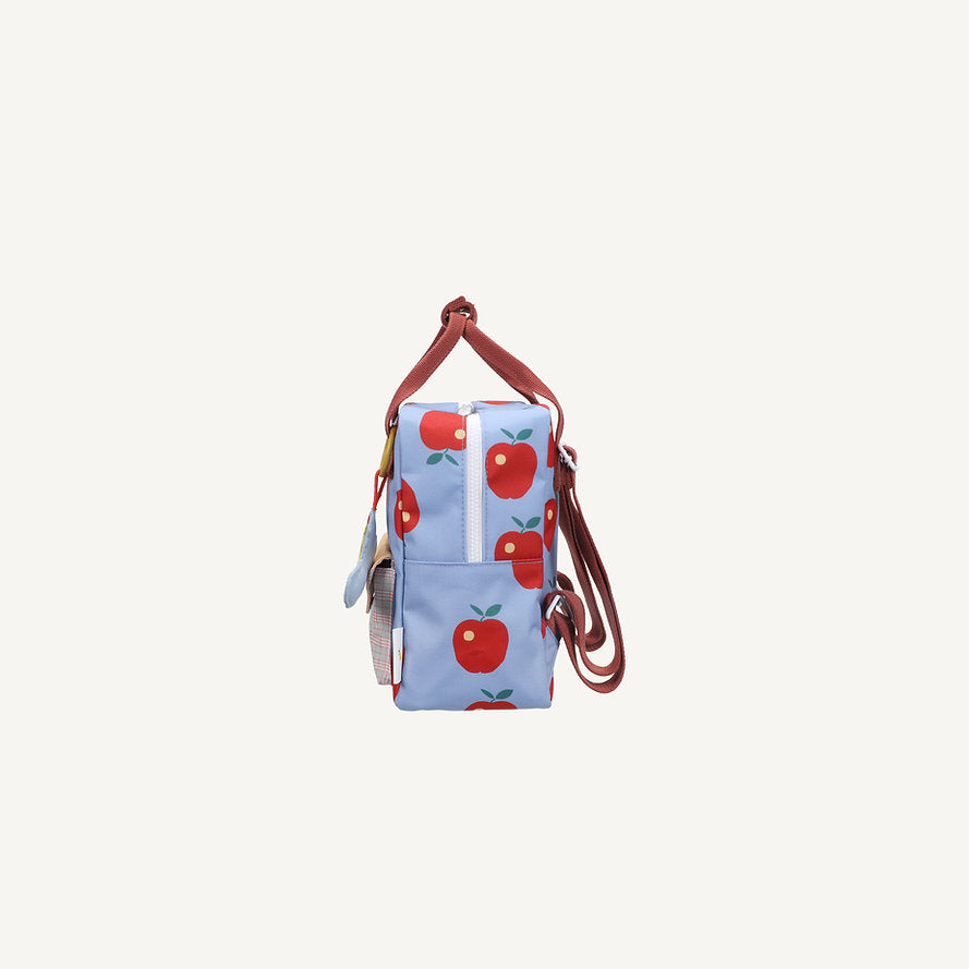 Side view of the Sticky Lemon Backpack Small - Special Edition - Apple Grub: a compact kids’ backpack with red apple prints, brown handles and straps, zipper closure, and made from recycled PET bottles against a light blue background.
