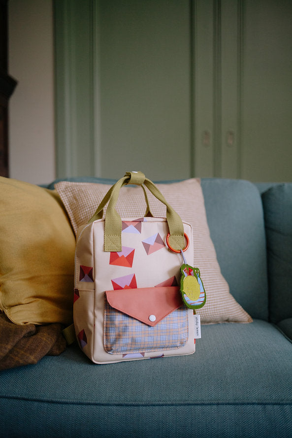 The Sticky Lemon Backpack Small - Special Edition - Snail Mail sits on a blue sofa with beige and yellow cushions. This kids' backpack has green handles, a front pocket, and is made from recycled PET bottles. Includes a green and yellow character keychain.