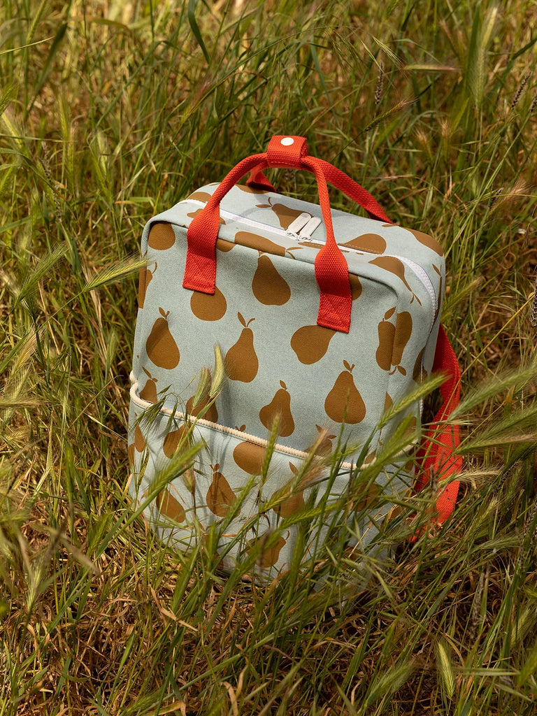 A Sticky Lemon Backpack Large with a pear print and red handles sitting in tall green grass, featuring a reliable YKK zipper for secure closure.